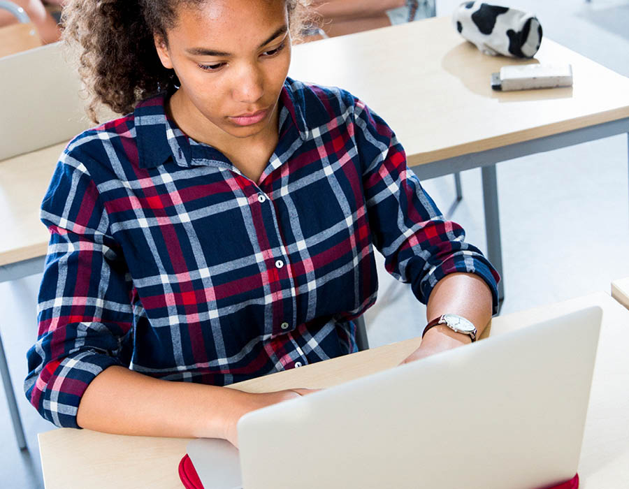 Flicka i mörkt hår och rödblårutig skjorta knappar på en dator i skolmiljö.